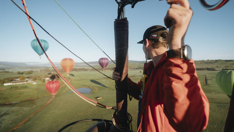 A man in a red jacket and black cap looks at four hot air balloons in the sky from his own hot air balloon basket