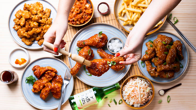 A table with several plates of fried chicken, fries and coleslaw. A woman standing over the table brushing sauce onto a piece of chicken.