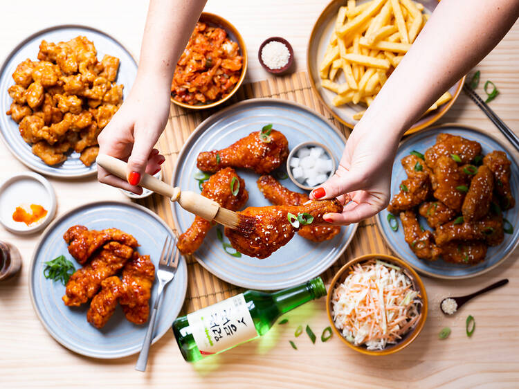 A table with several plates of fried chicken, fries and coleslaw. A woman standing over the table brushing sauce onto a piece of chicken.