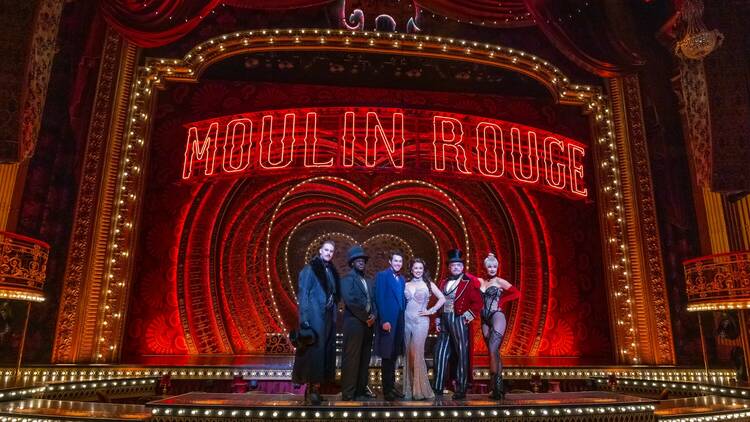 A group of six performers stand in front of a red theatre stage that says Moulin Rouge