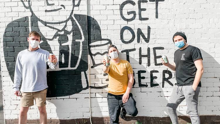 Three people standing in front of a white brick wall that has a painting of Dan Andrews and text that says 'Get on the beers'.