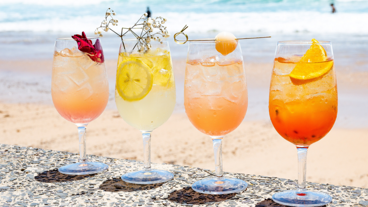 Four cocktails in wine glasses lined up in front of the ocean