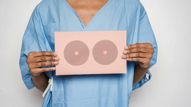 Woman in blue medical gown holds a card of illustrated breasts in front of her chest.
