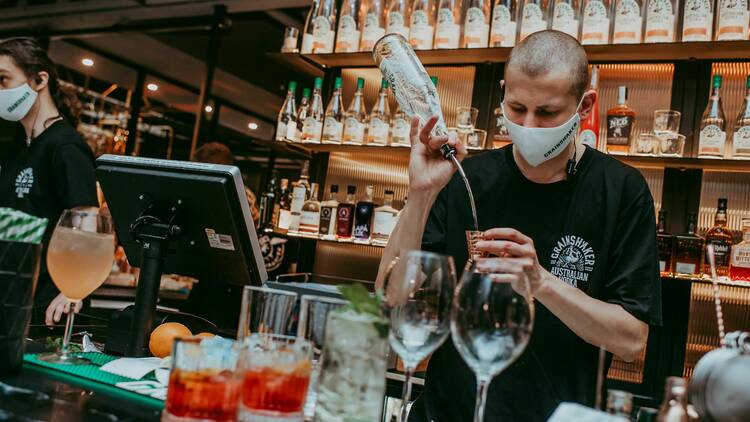 A masked bartender pouring drinks behind the bar at the Distiller.