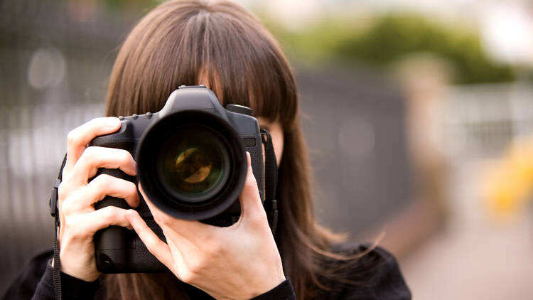 A brunette woman holds a large black camera up to her face to take a photo.