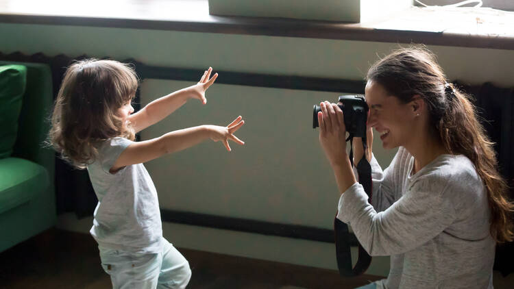 A woman crouching down takes a photo of a small child with outstretched arms.