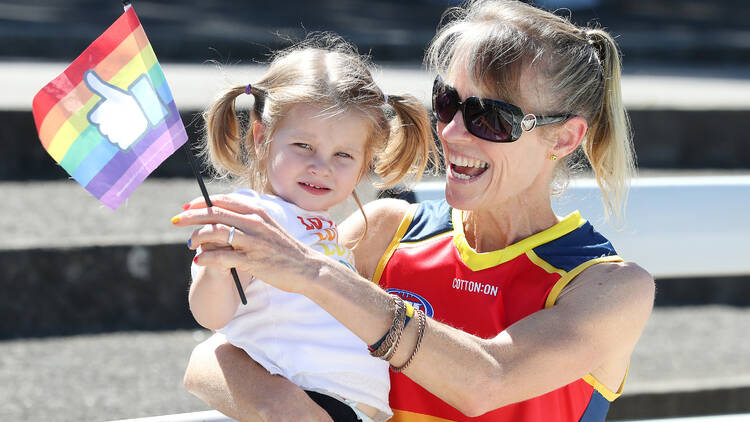 A woman in a rainbow jersey holding a small child while waving a rainbow flag.