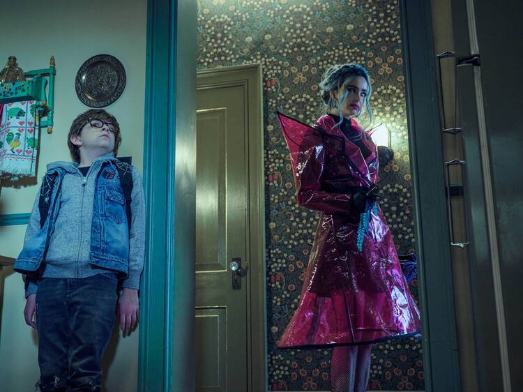  A still from the Netflix Original movie Nightbooks. A young boy hides from a woman wearing a pink coat. 