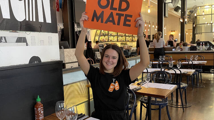 A woman in a black t-shirt holds an orange sign up in a restaurant that says 'Old Mate'