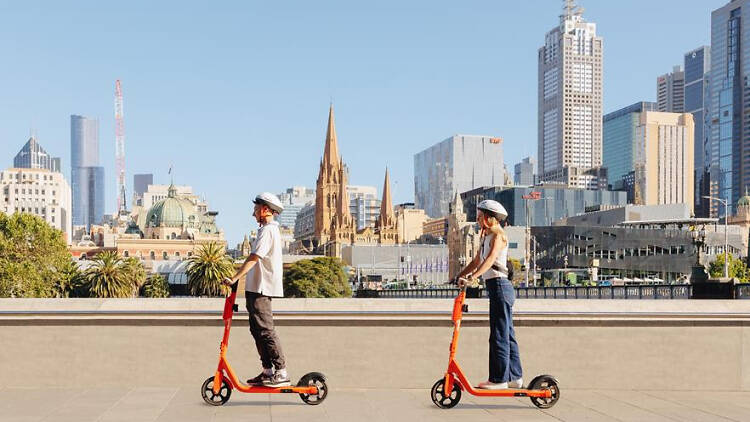 Two people riding bright orange e-scooters in Melbourne, with the city skyline in the background