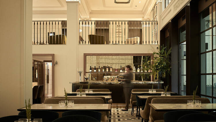 A waiter making their way through Luci restaurant, a room filled with marbled tabletops and plush chairs.