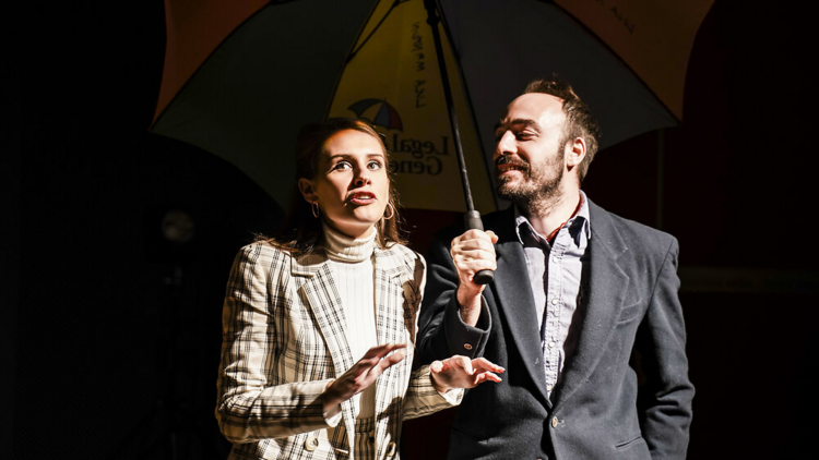 A man in a grey suits holds an umbrella up for a woman in a check-print jacket