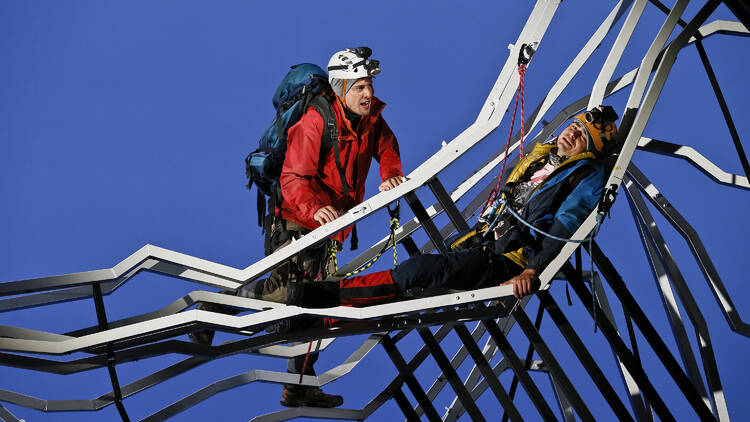 A man lies ona mountain made of steel girders while another man in climbing equipment seemingly finds him