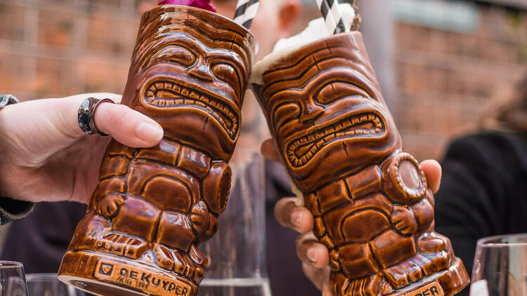 Two brown tiki mugs are lifted together in a 'cheers' motion