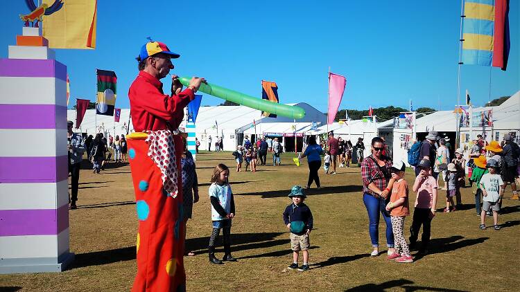 A festival ground surrounded by white marquees. Adults and children are throughout the space, with a clown on stilts blowing up a long balloon.