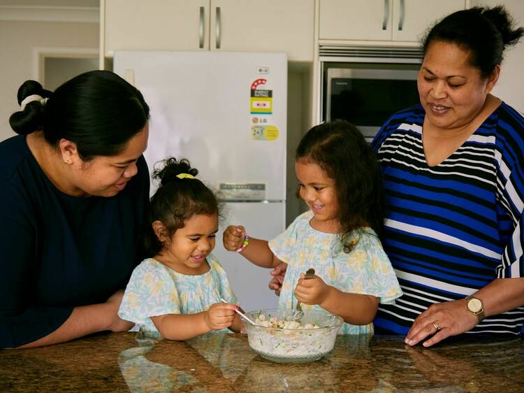 Sela Atiola stands to the left of her kitchen bench with her daughters in the centre and her mother to the far right. They make 'ota ika together with smiles on their faces.