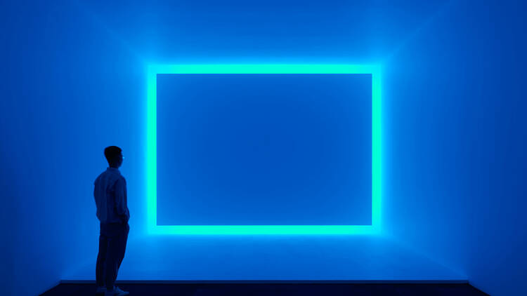 A large, blue, neon rectangle glowing against a blue wall. A person stands admiring it and showing that the rectangle is at least as tall as a man