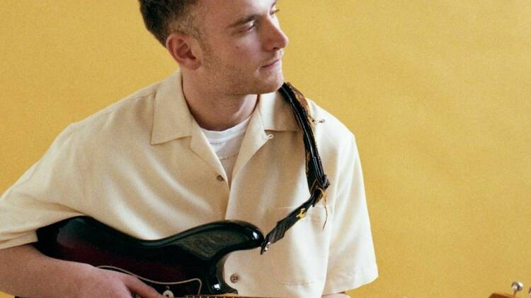 Musician Tom Misch holding a guitar and sitting against a yellow backdrop.