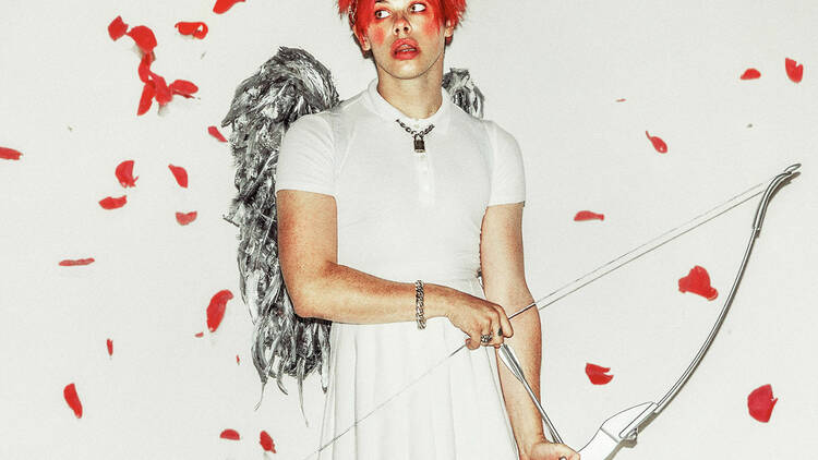 Musician Yungblud wearing a white top and skirt, a pair of silver wings and holding a bow and arrow.