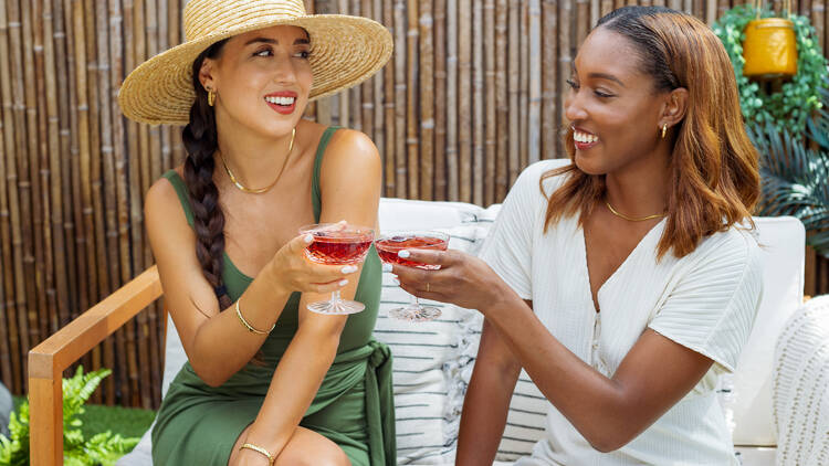 Two women sitting down cheers with their cocktails.