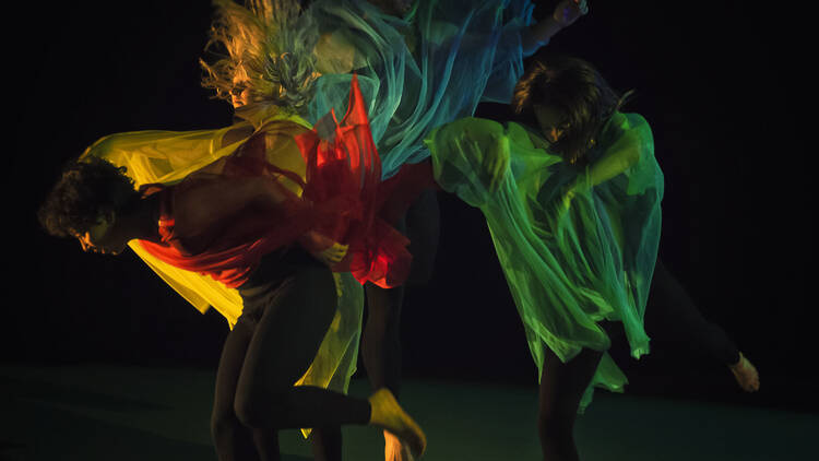 Three dancers in a dark space moving with transparent, coloured shrouds over their arms and shoulders