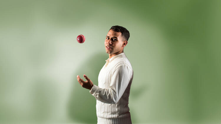 A man wearing cricket whites smiles slightly as he tosses a cricket ball in the air