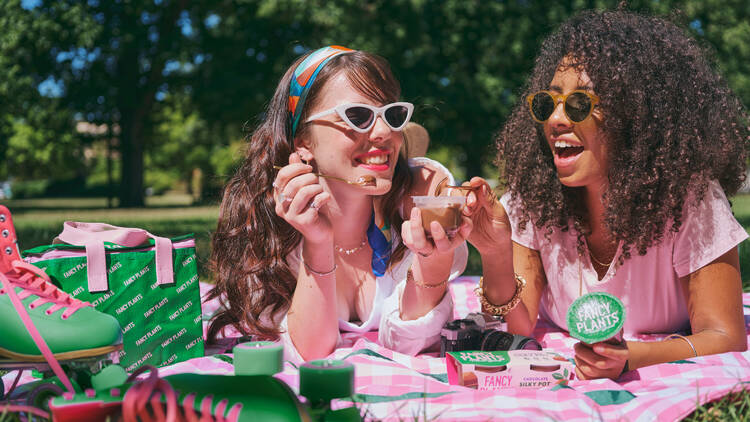 Two girls with curly brown hair lay on a picnic rug eating chocolate pudding.