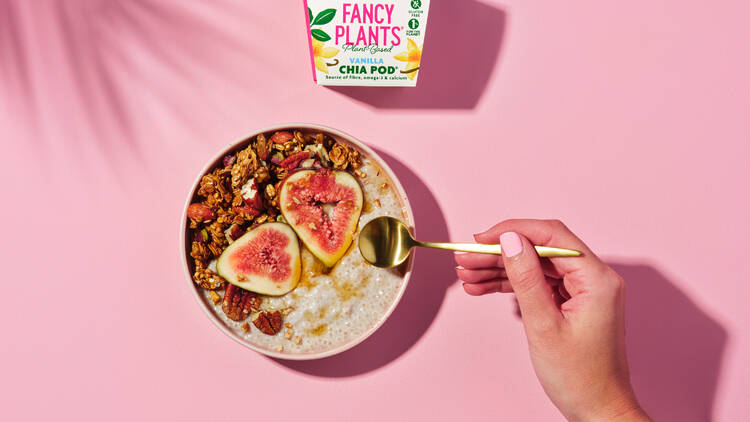 A person holds a gold spoon out to a bowl of cereal, fruit and chia pudding against a pink backdrop.