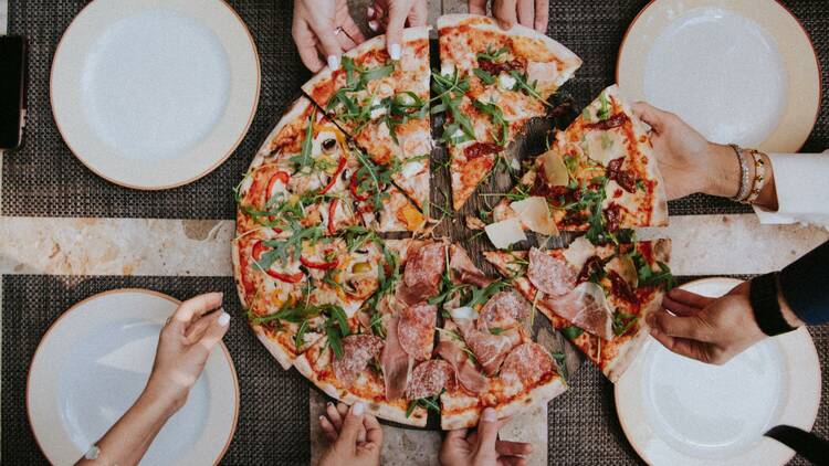 An overhead shot of lots of hands reaching for a slice of pizza.