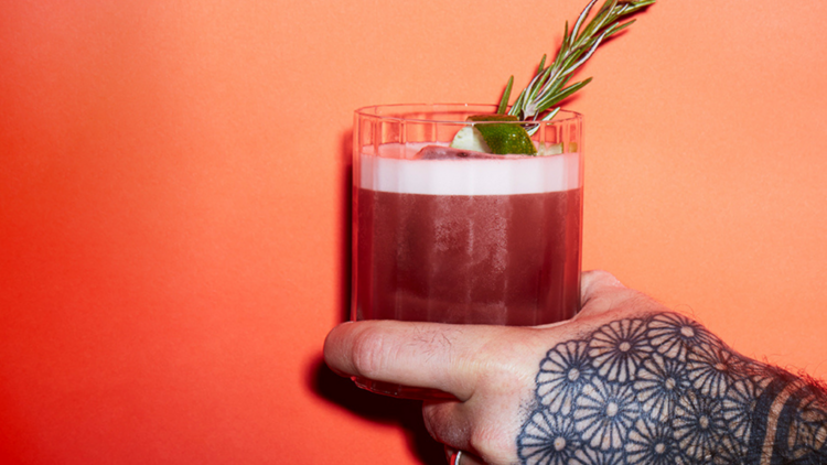 A tattood hand holding a cocktail in front of a pink wall