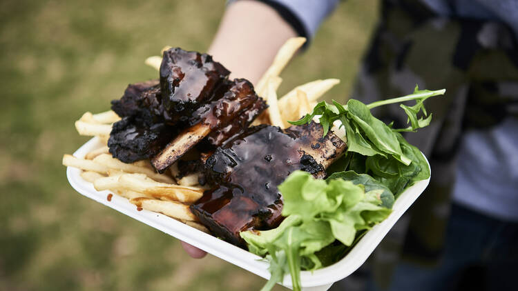 A person holds out a BBQ sauce-laden pack of ribs, chips with a handful of fresh greens stuffed in the side.