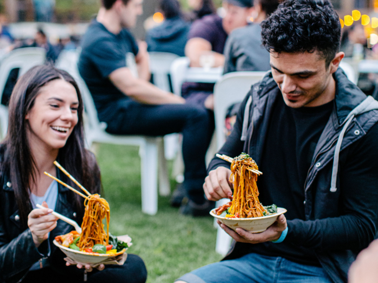 Two people sit on the grass eating noodles and smiling