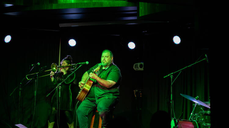 A man bathed in a green light sits on stage in front of a microphone with a guitar.