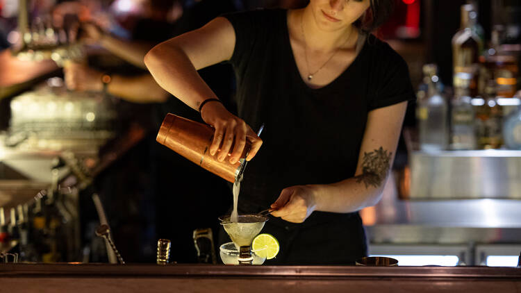 A woman at a bar pours a cocktail into a Martini glass.
