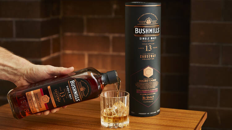 Bushmills Causeway Collection Dinner A person pours Bushmills Irish Whiskey into a glass with ice on a wooden table