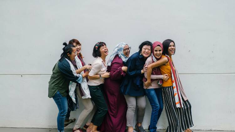A group of women standing against a wall, laughing and holding each other.