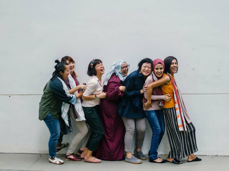 A group of women standing against a wall, laughing and holding each other.