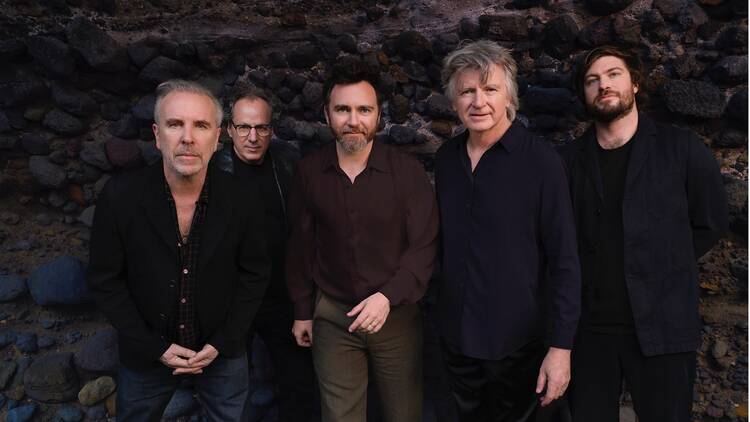 The band Crowded House standing in front of a rocky stone wall.