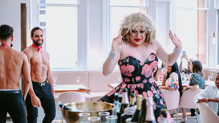 A drag queen in the middle of a sunny room with soft pink furnishings. Two male topless waiters look on smiling in the background.