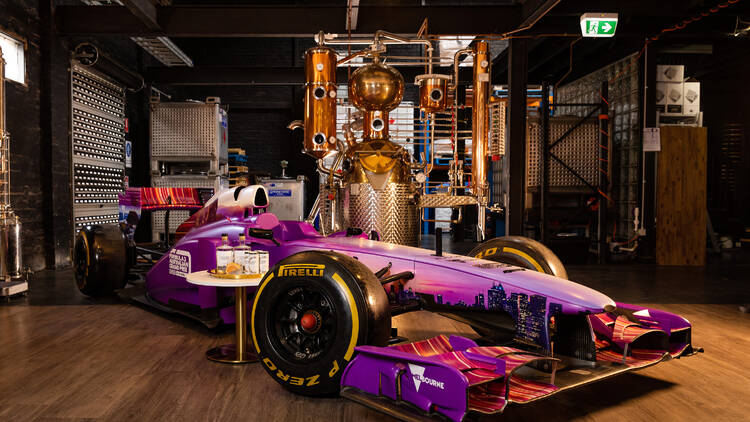 A purple F1 car parked in a distillery next to a large copper still and a table with some Patient Wolf gin bottles on it.