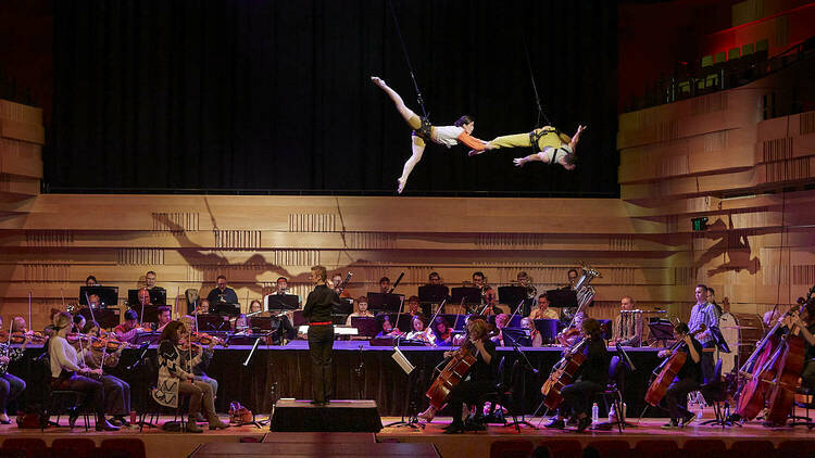 Two aerialists perform a high-flying routine while suspended over an orchestra.