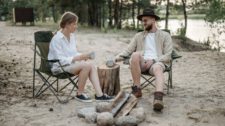 A man and a woman sitting by a campfire and enjoying a cup of coffee.
