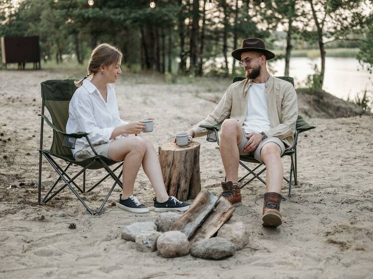 A man and a woman sitting by a campfire and enjoying a cup of coffee.