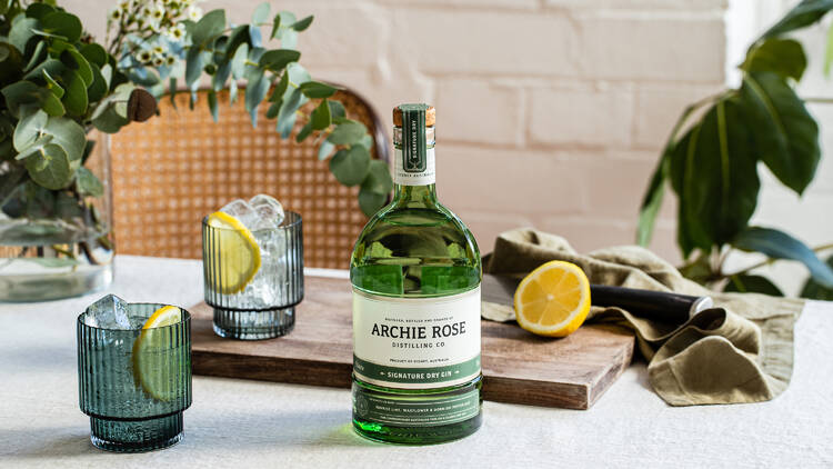 A bottle of Archie Rose Distilling Co gin on a table with glasses, half a lemon and some greenery.