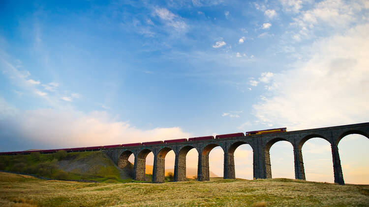 Train crosses the famous Ribblehead Viaduct. The Ribblehead Viaduct or Batty Moss Viaduct carries the Settleâ€“Carlisle Railway across Batty Moss in the valley of the River Ribble at Ribblehead.