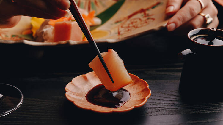 A piece of raw fish is being dipped into a small flower-shaped dish filled with soy sauce.