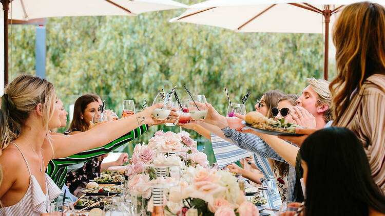 A group of friends sitting at a table and cheersing with cocktails and wine.