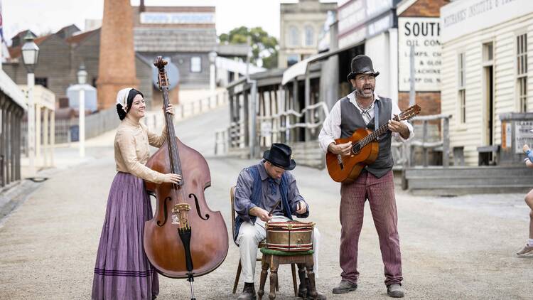 Three musicians dressed in old-fashioned clothes play their instruments on a historic-looking street.