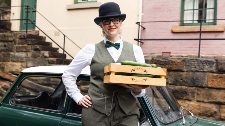 A woman in old timey clothes holding a crate of gin and cucumbers