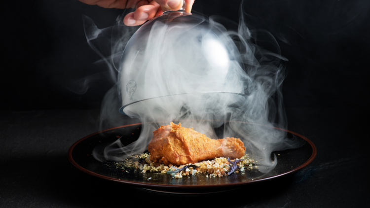 A fried chicken drumstick surrounded by smoke under a cloche